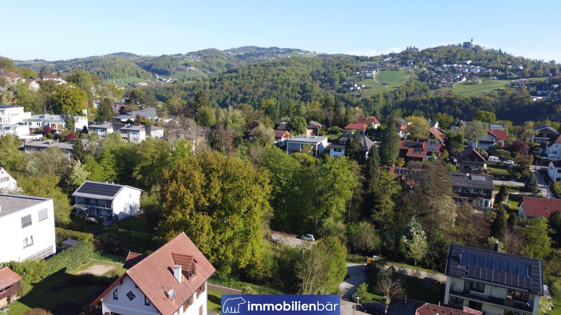 Panoramablick aus der Luft auf eine grüne Wohngegend mit bewaldeten Hügeln am Horizont.