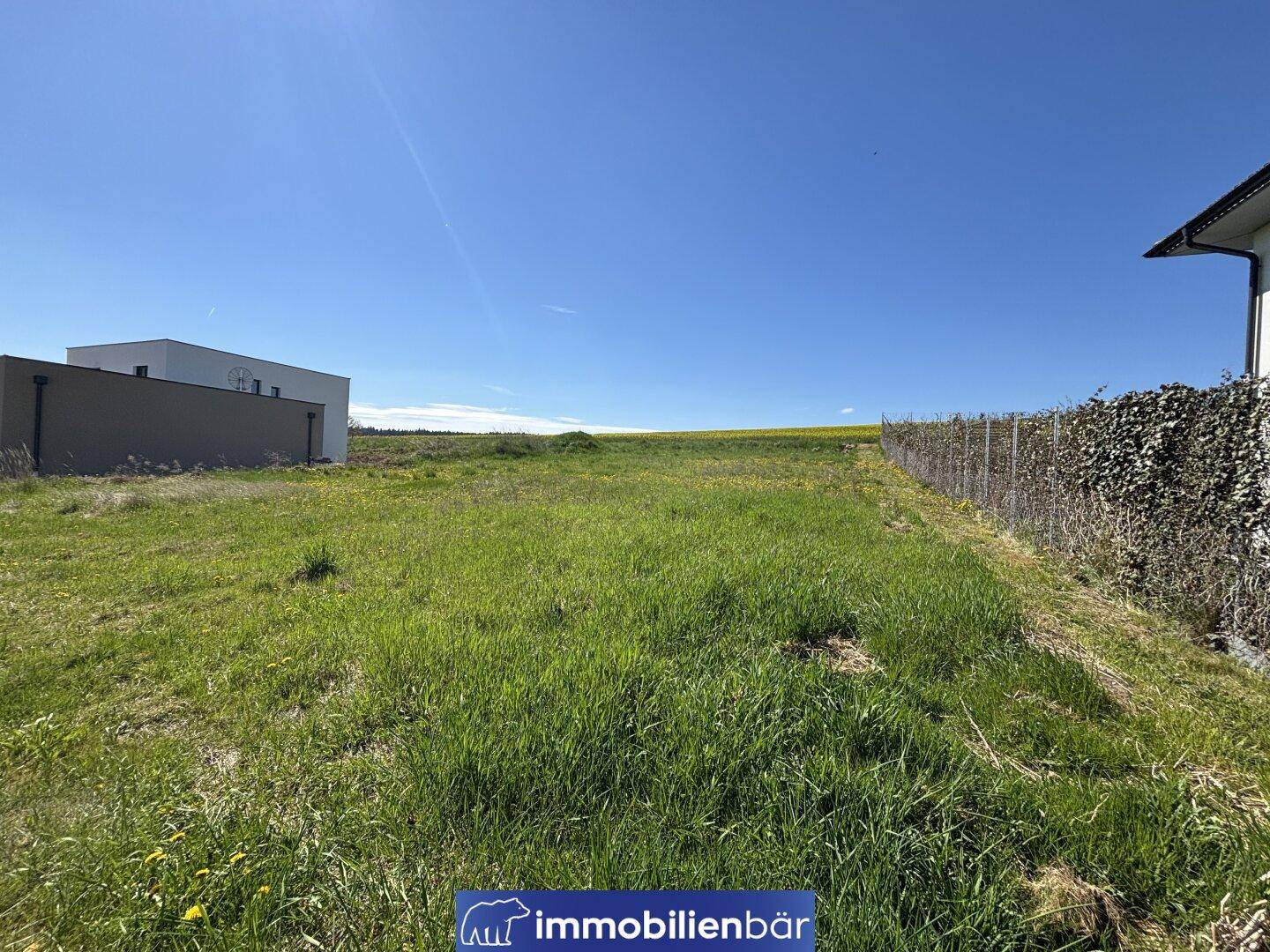 Blick über das grüne Baugrundstück mit weitem Horizont und angrenzenden Feldern unter blauem Himmel.