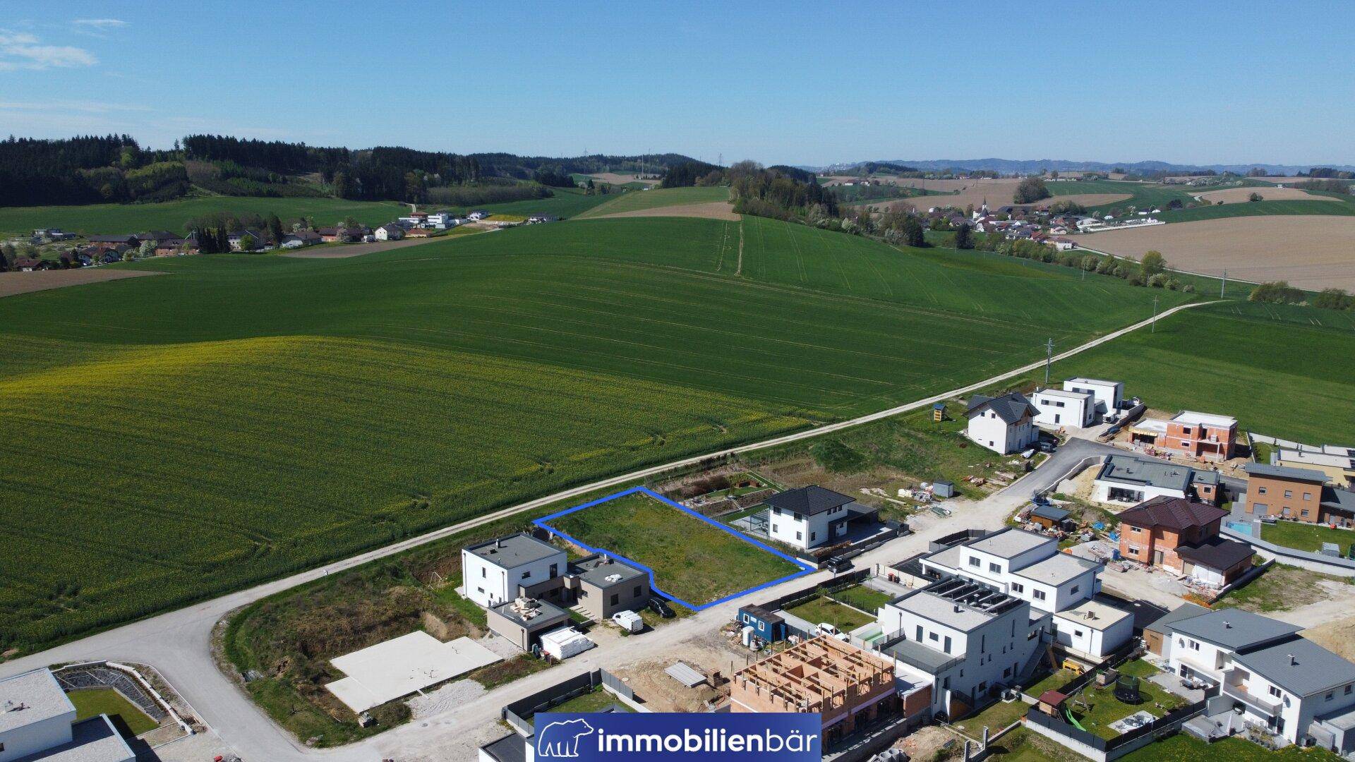 Weitläufige Luftaufnahme des Baugrundstücks, umgeben von Feldern und einer wachsenden Wohnsiedlung.