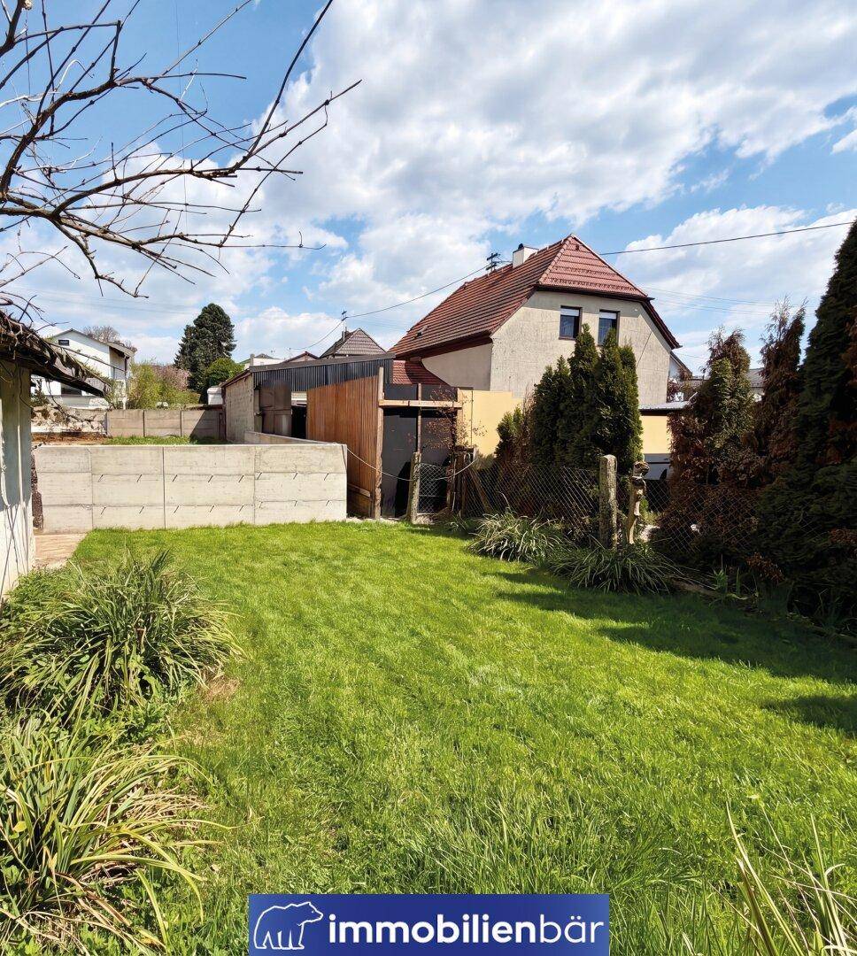 Grüner Hinterhof mit Rasenfläche, umgeben von einem Zaun und einem Wohnhaus im Hintergrund.