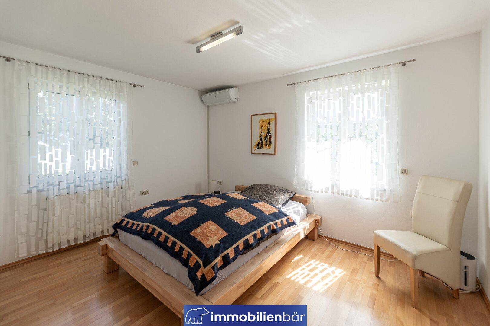 Helles Schlafzimmer mit Holzbett, Klimaanlage und zwei Fenstern für viel Tageslicht.