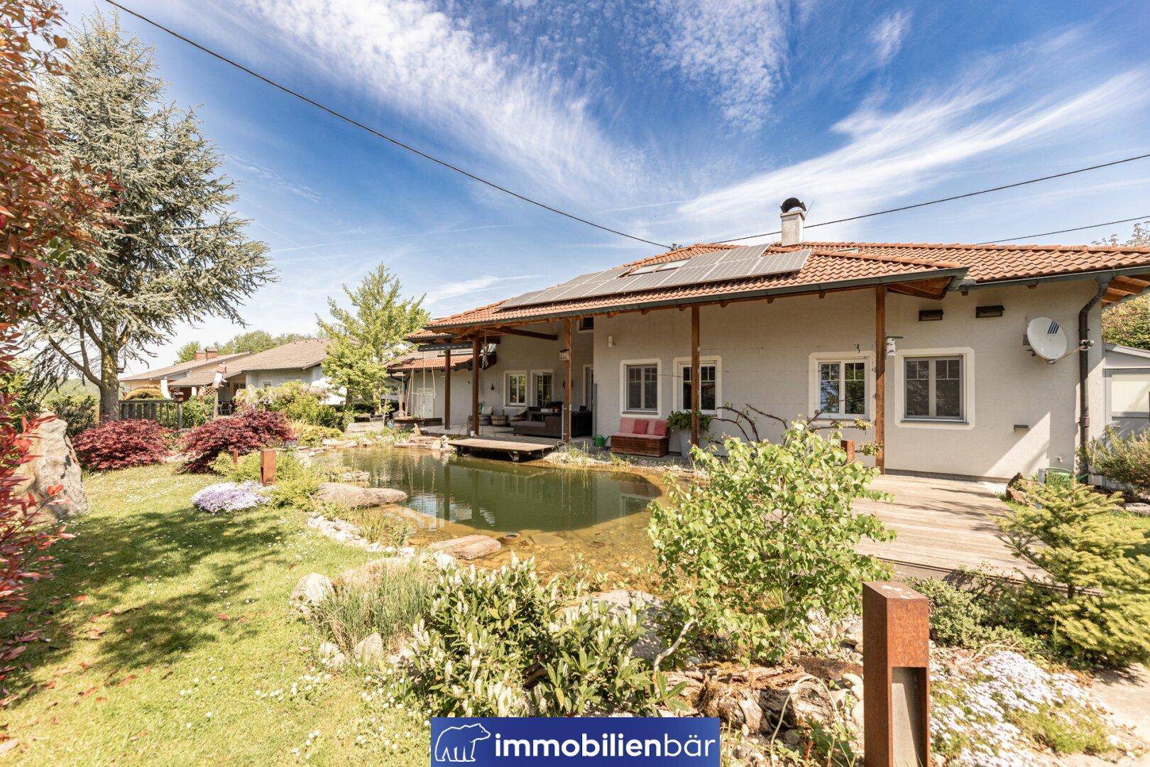 Rückansicht des Hauses mit überdachter Terrasse, Teich und Photovoltaikanlage auf dem Dach.
