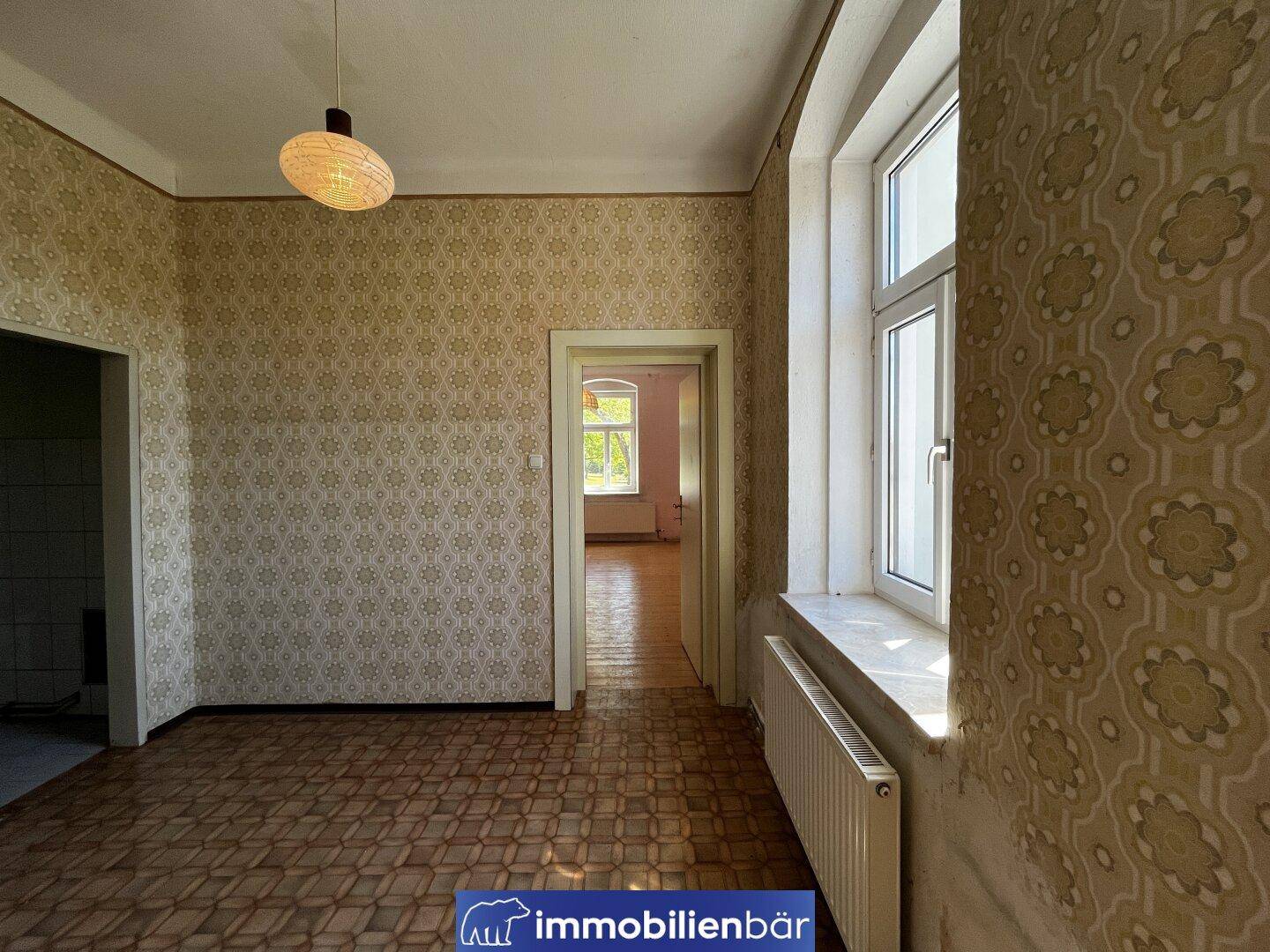 Zimmer mit gemusterten Tapeten, Linoleumboden und einem großen Fenster für Tageslicht.