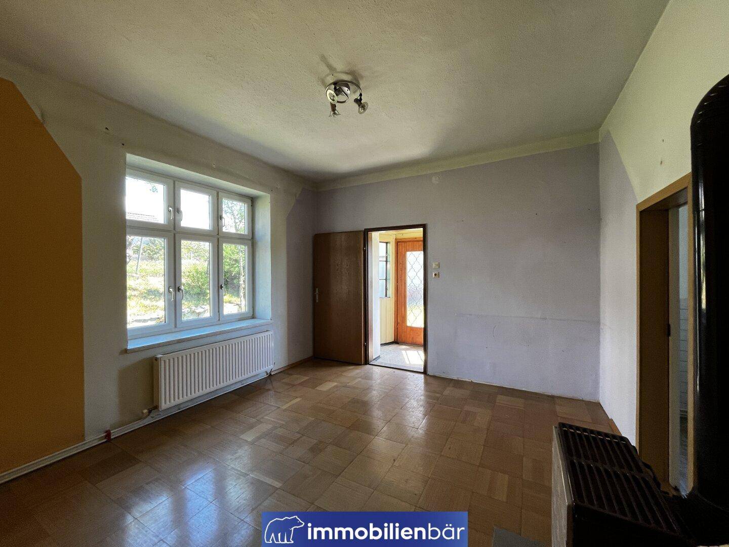 Helles Zimmer mit Parkettboden, Kachelofen und einem großen Fenster.