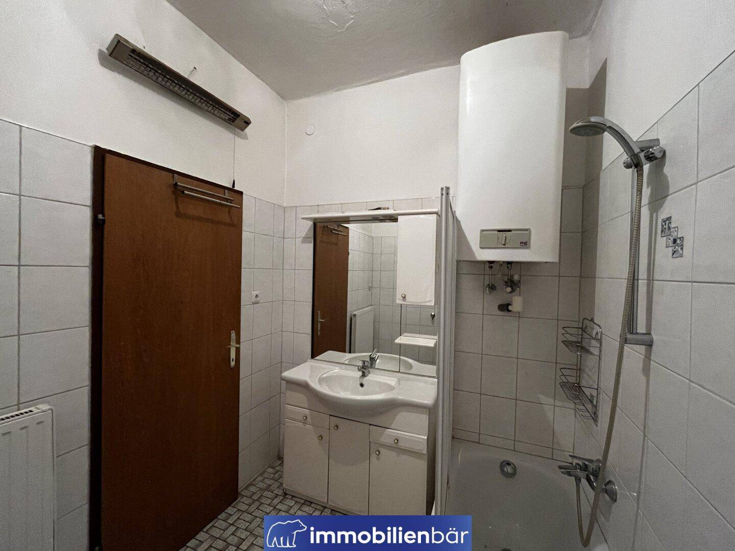 Kleines Badezimmer mit Dusche, Waschbecken und Boiler, gefliest in Weiß.