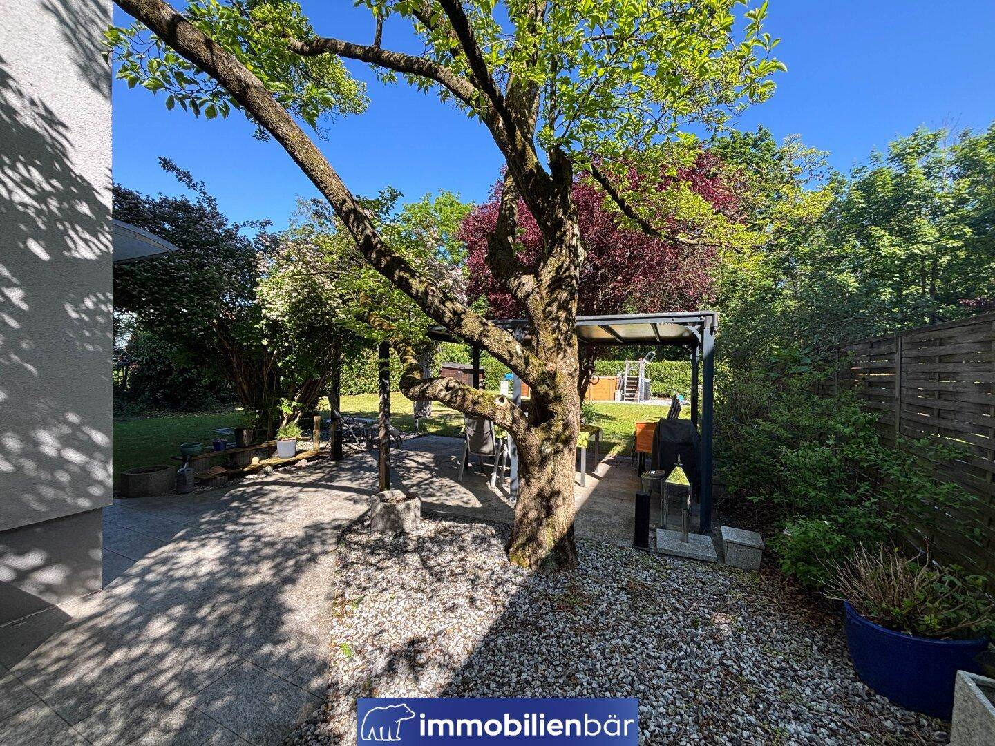 Gepflasterte Terrasse mit einem großen Baum, überdachtem Sitzbereich und Zugang zum Garten.