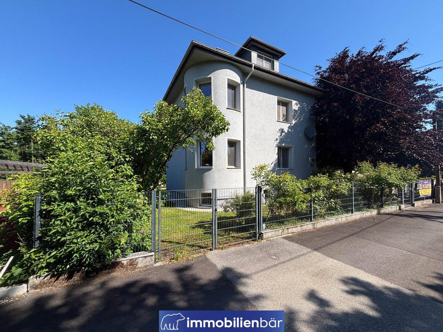 Ansicht des Hauses von der Straße mit Zaun, Bäumen und einer hellen Fassade.