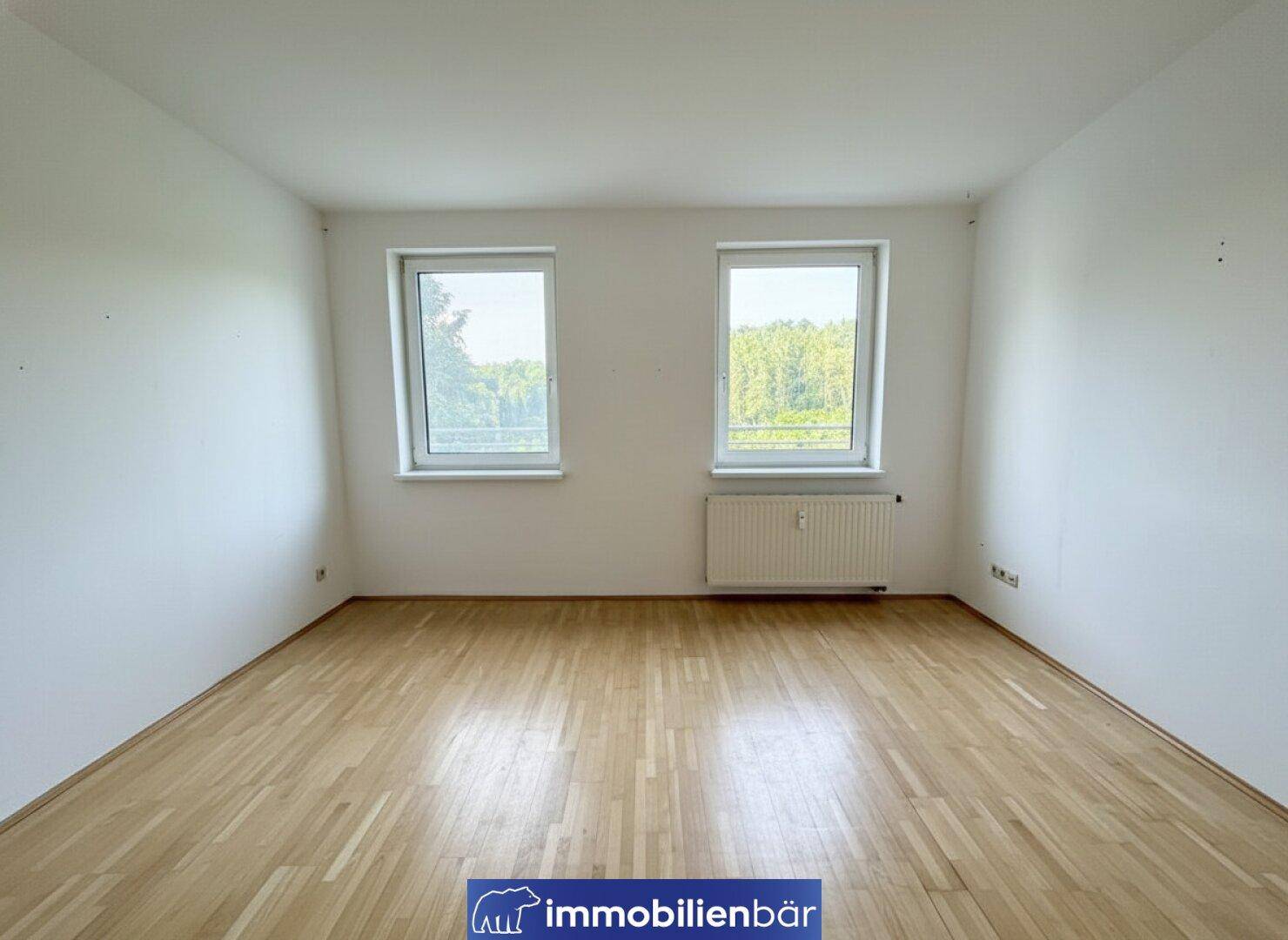 Helles Zimmer mit Parkettboden, weißen Wänden und Blick ins Grüne durch zwei Fenster.