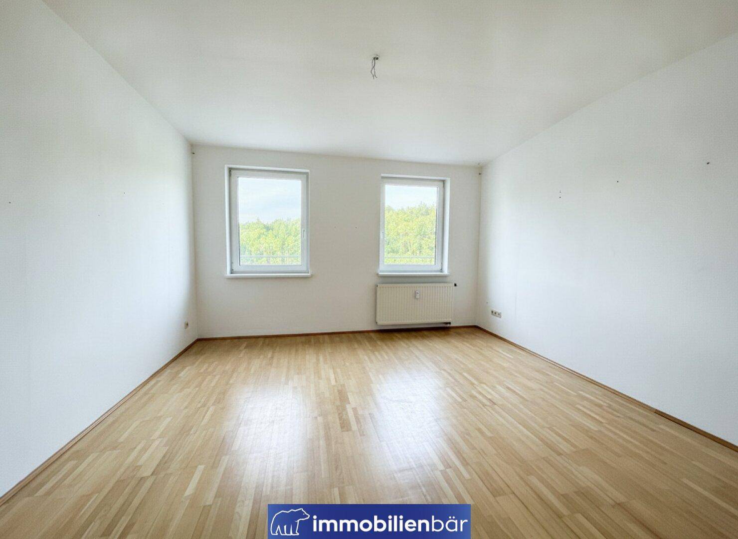 Geräumiges Zimmer mit hellem Parkettboden und zwei Fenstern, die viel Tageslicht hereinlassen.