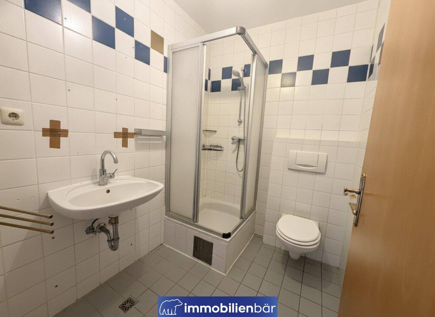 Badezimmer mit Duschkabine, Waschbecken und WC, gefliest in Weiß mit blauen und goldenen Akzenten.