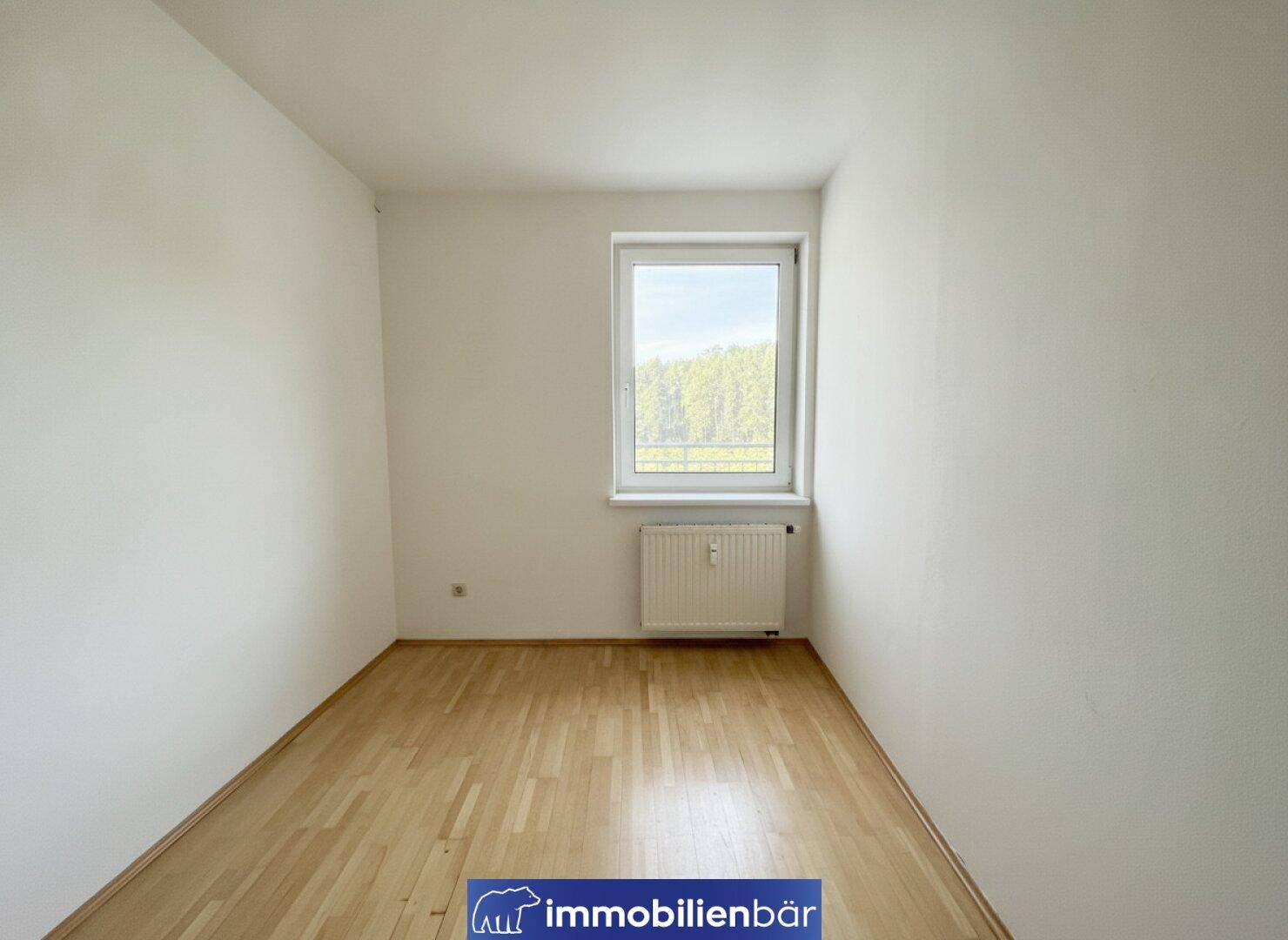 Kleineres Zimmer mit Parkettboden, weiß gestrichenen Wänden und einem Fenster mit Blick ins Grüne.
