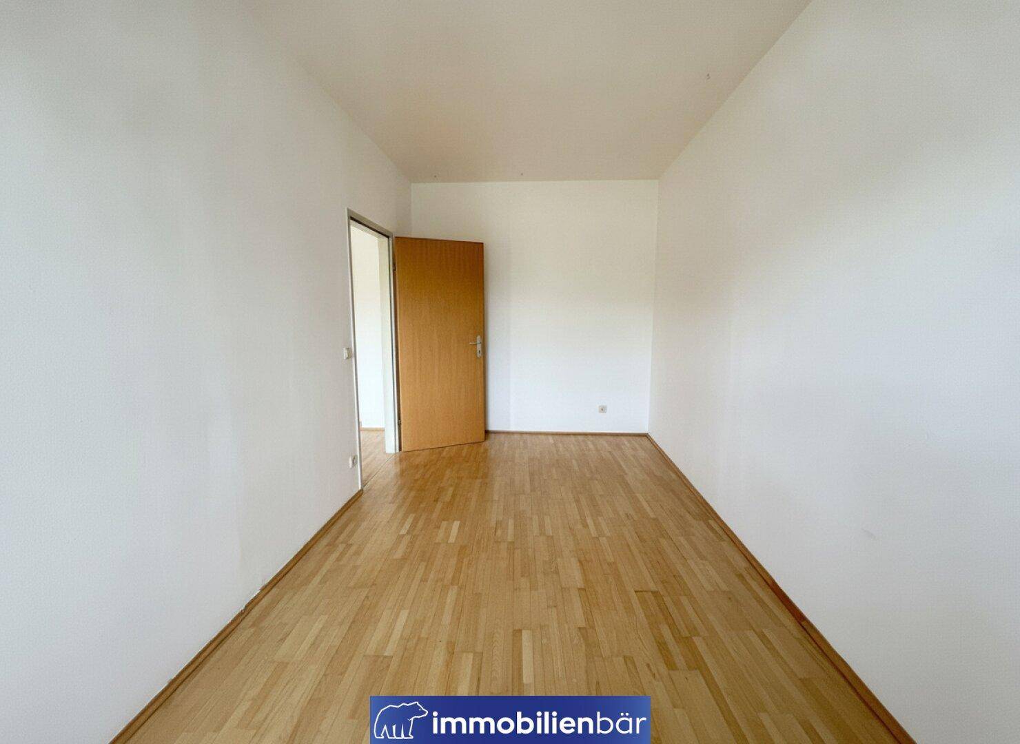 Leeres Zimmer mit Parkettboden und weiß gestrichenen Wänden, eine offene Tür führt in den Flur.