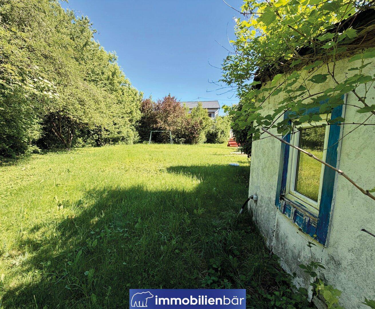 Sonniger Gartenbereich mit einer kleinen Hütte und üppiger Vegetation, umgeben von Bäumen.