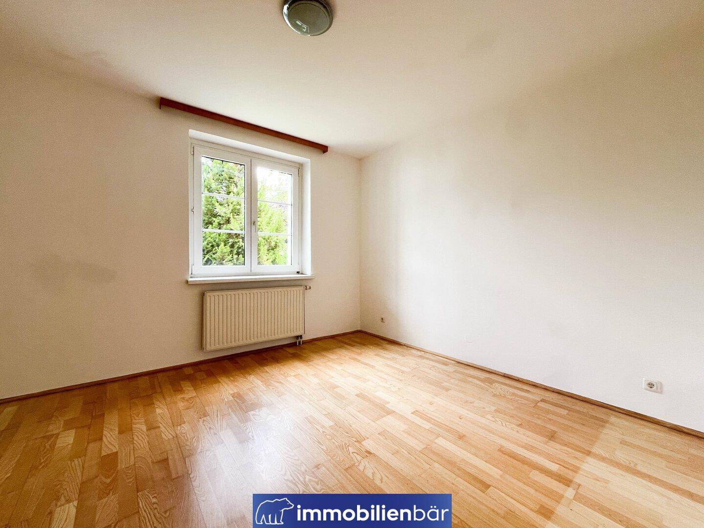 Helles Schlafzimmer mit Parkettboden und großem Fenster mit Grünblick.