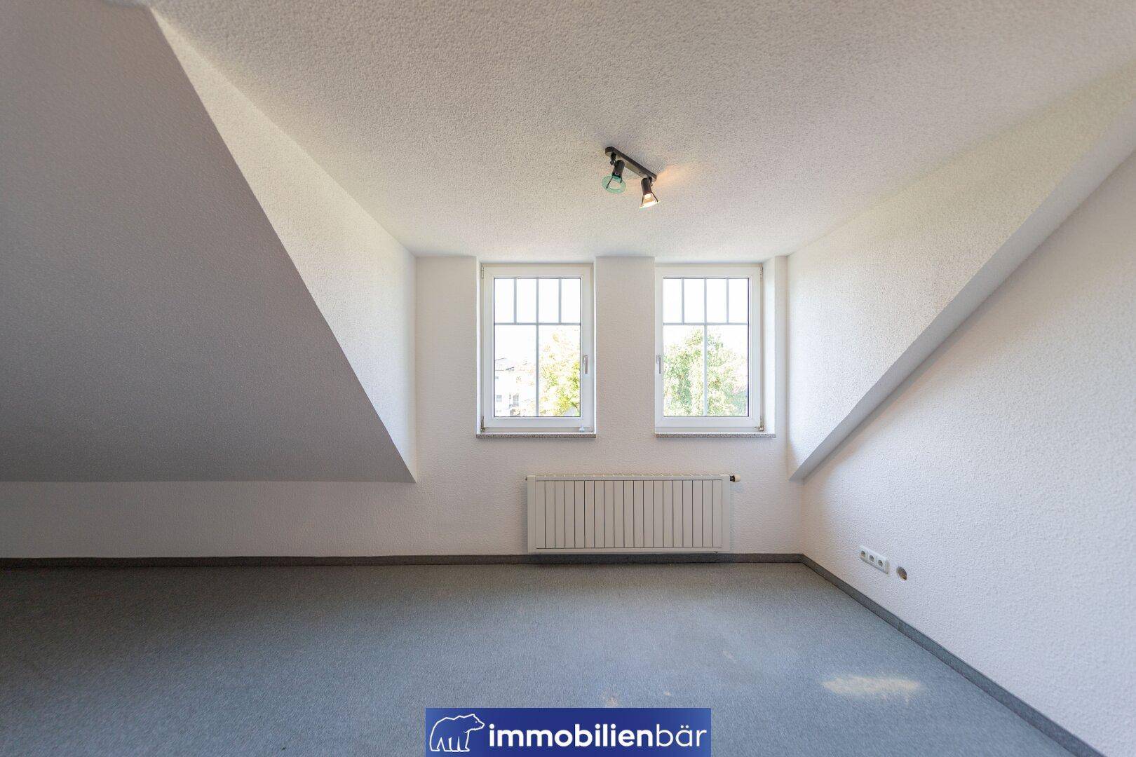 Geräumiges Zimmer im Dachgeschoss mit zwei Fenstern, Schrägen und Teppichboden, gut beleuchtet.