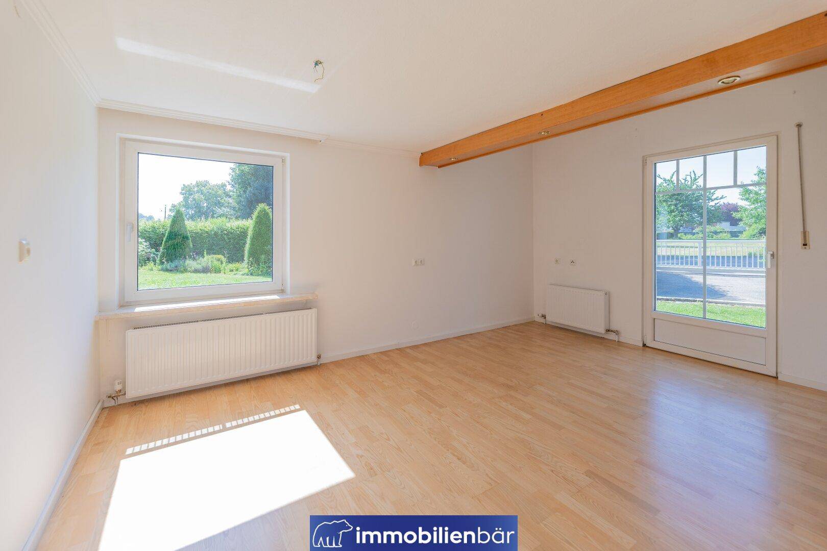 Geräumiger, heller Raum mit Holzboden, großem Fenster und Holzbalken an der Decke.