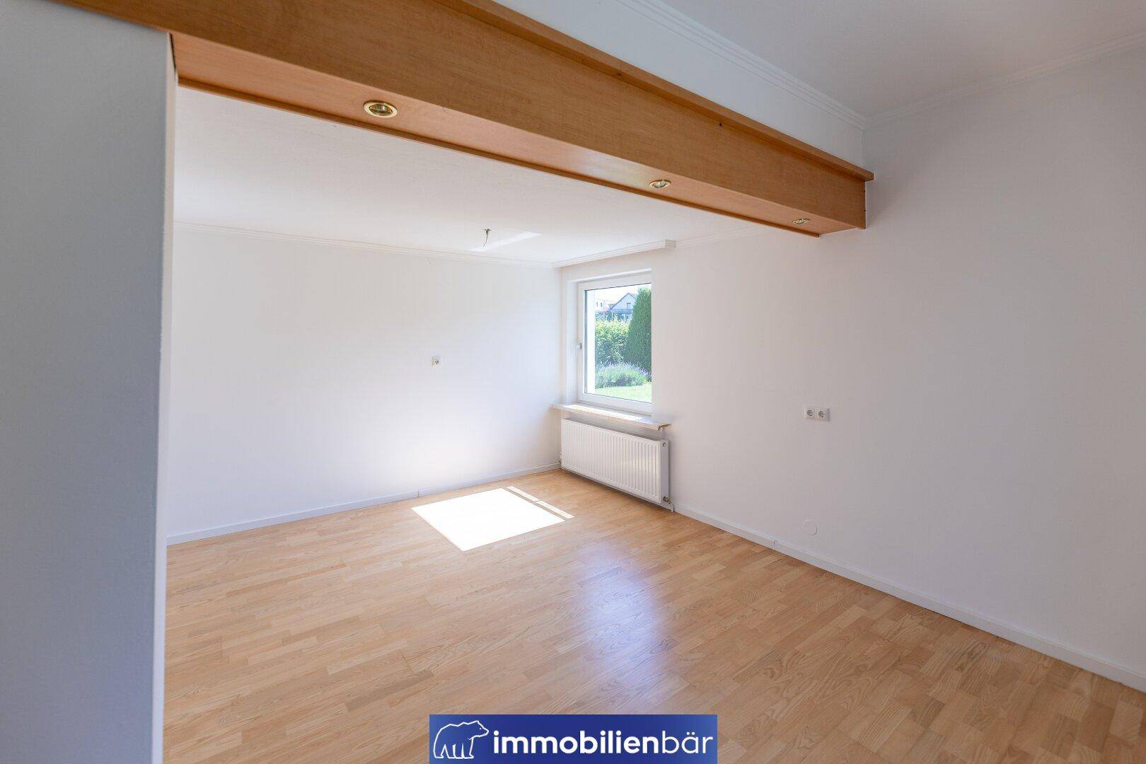 Heller Raum mit Holzboden, großem Fenster und einem dekorativen Holzbalken an der Decke.