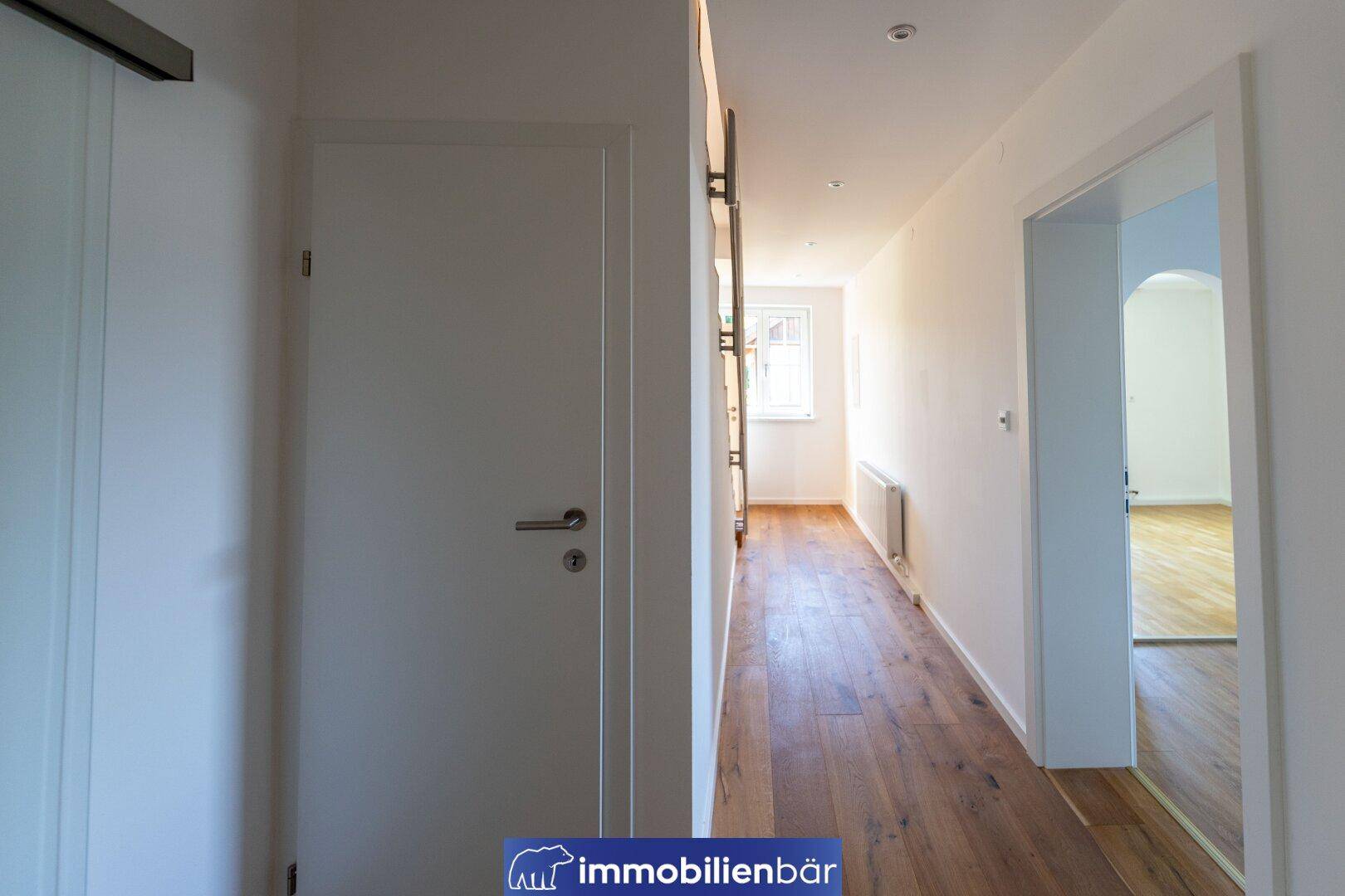 Langer, heller Flur mit Holzboden, weißen Türen und einem Fenster am Ende des Ganges.