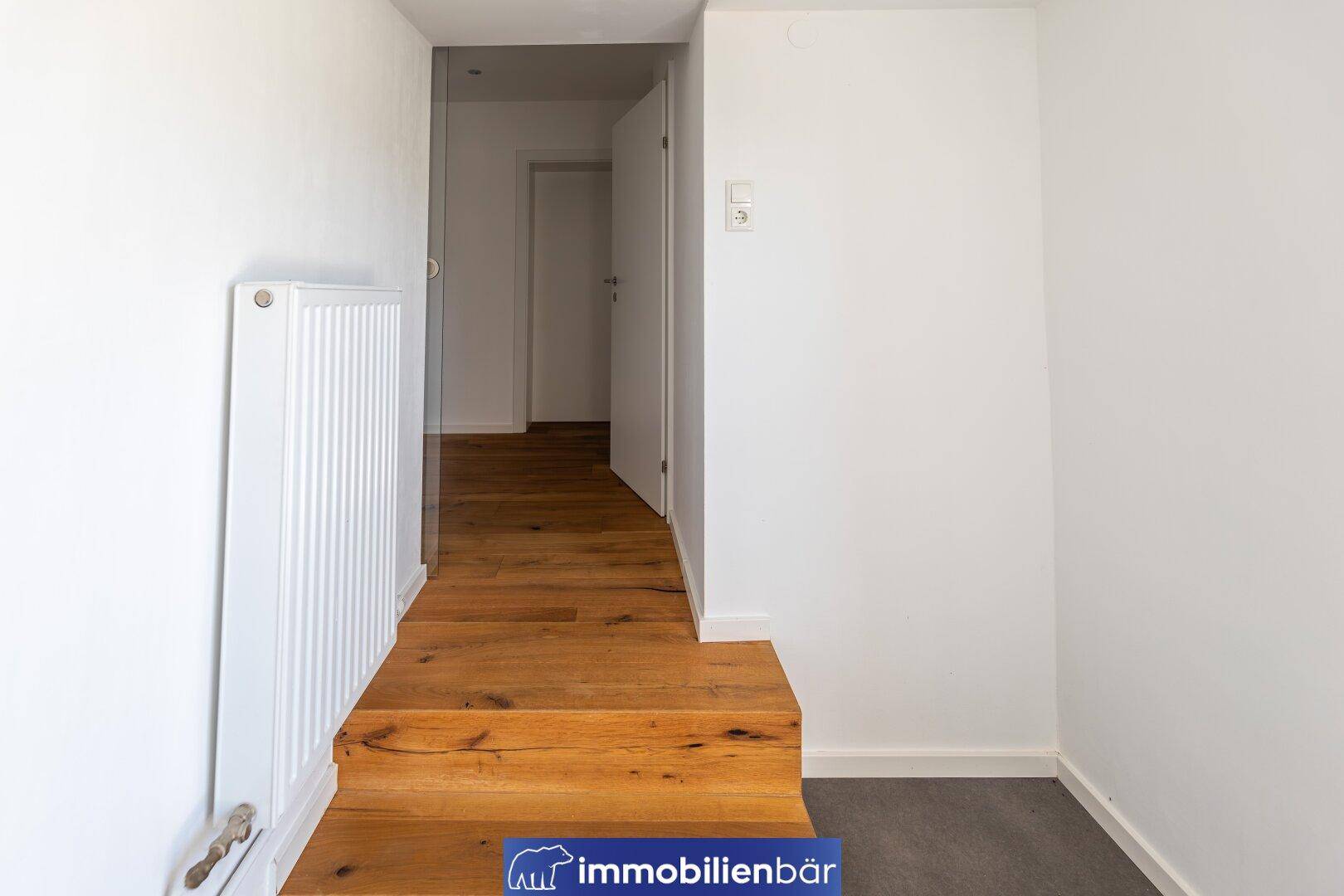 Heller Flur mit Holztreppe, weiß gestrichenen Wänden und einem Heizkörper.