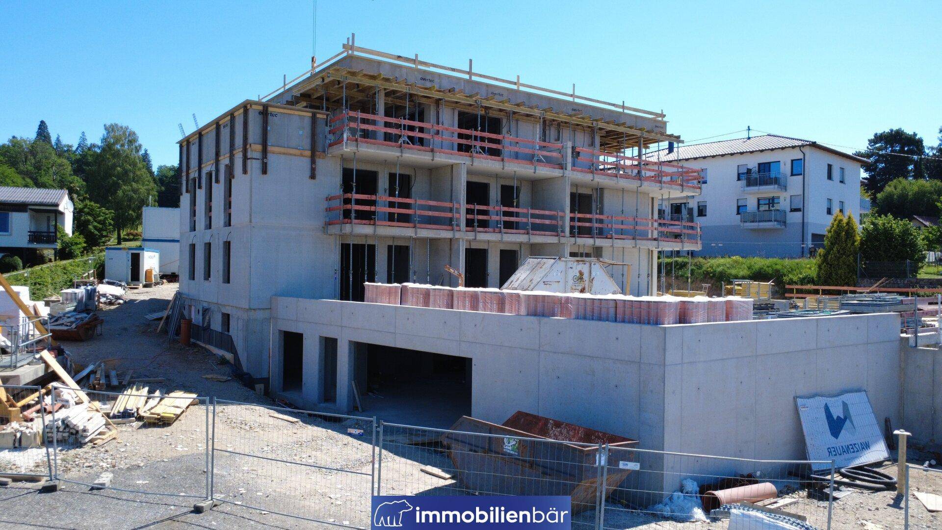 Neubau im Rohbau mit sichtbaren Balkonstrukturen, Baugerüst und Eingang zur Tiefgarage auf einer sonnigen Baustelle.