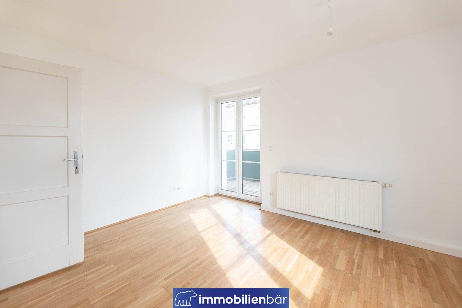 Helles Zimmer mit Parkettboden, weißen Wänden und Zugang zu einem Balkon.