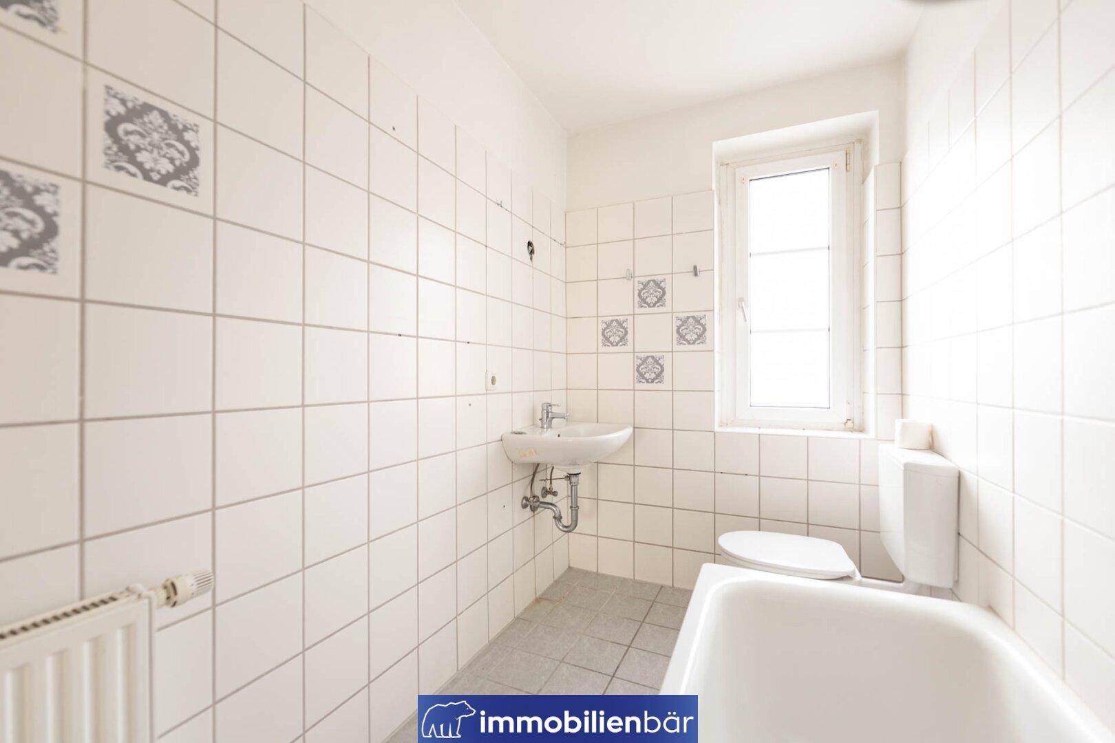 Badezimmer mit weißen Fliesen, Badewanne, Waschbecken, Toilette und Fenster.