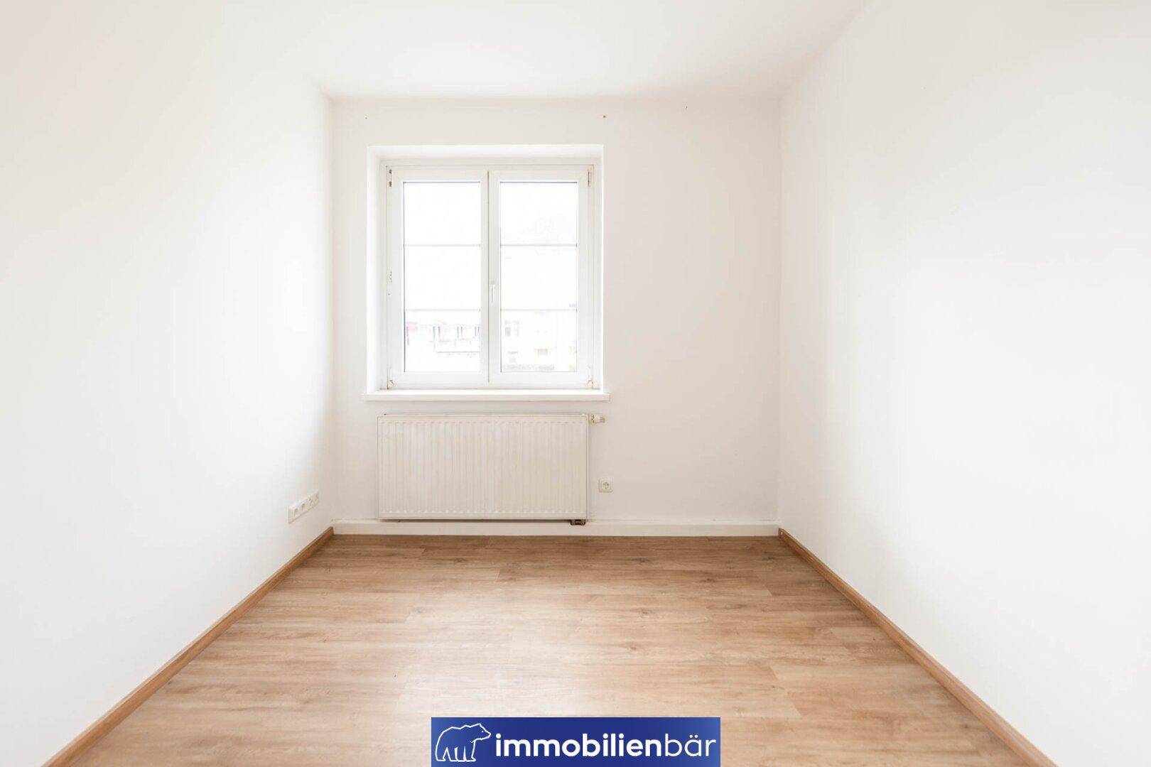 Leeres Zimmer mit Parkettboden, weißen Wänden und einem großen Fenster.