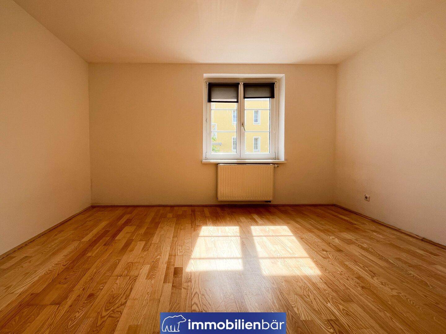 Helles Zimmer mit Holzparkettboden und einem Fenster, das Tageslicht hereinlässt.