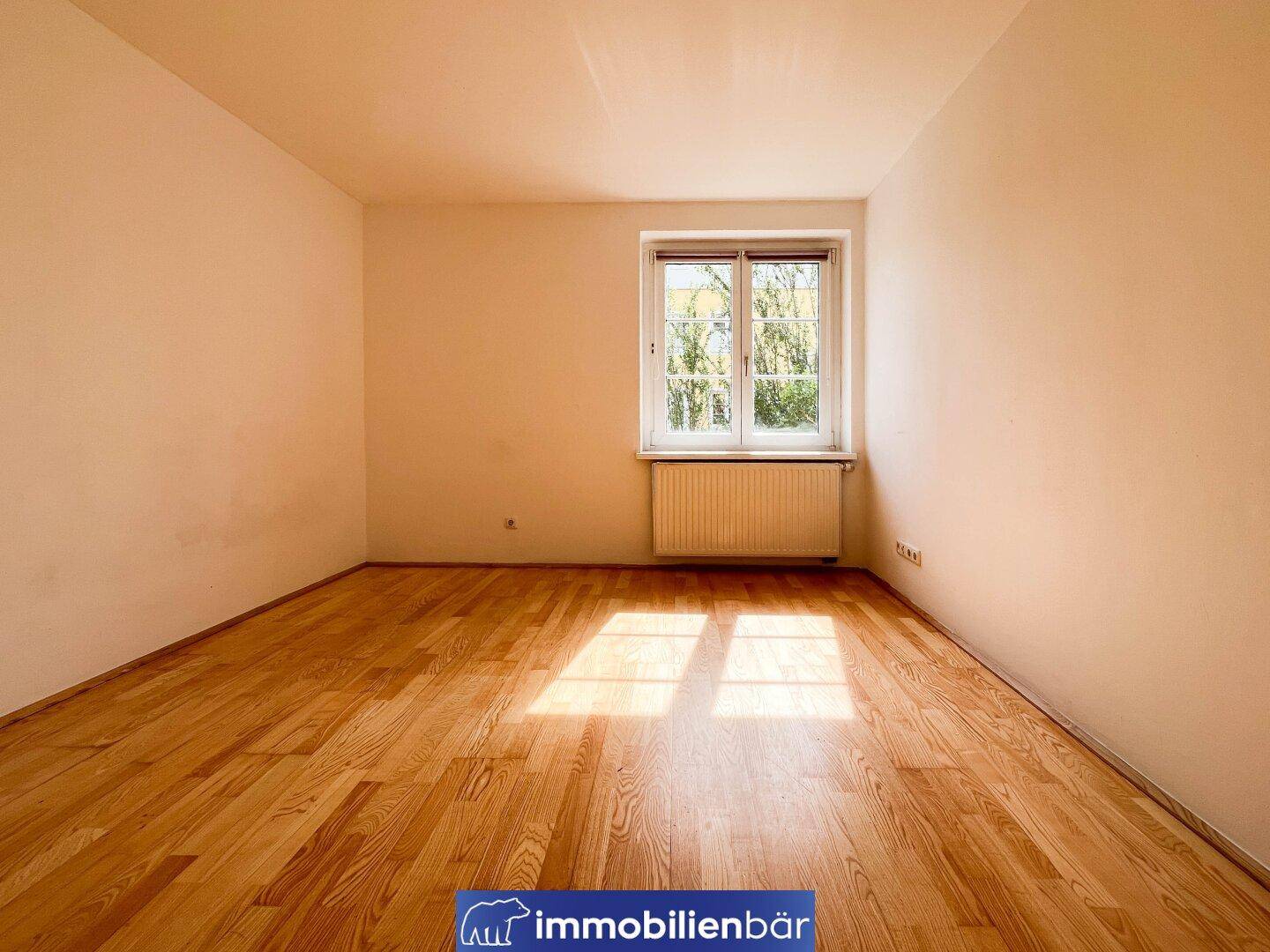 Geräumiges Zimmer mit Parkettboden und einem großen Fenster mit Blick ins Grüne.