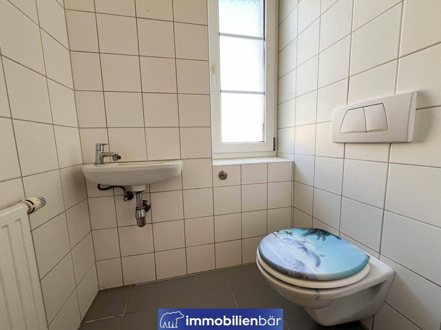 Kleines Gäste-WC mit Fenster, Waschbecken und Wand-WC mit dekorativem Deckel.
