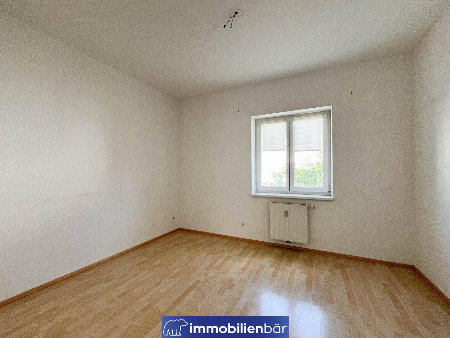 Ruhiges Schlafzimmer mit hellem Parkettboden und einem Fenster, das viel Tageslicht hereinlässt.