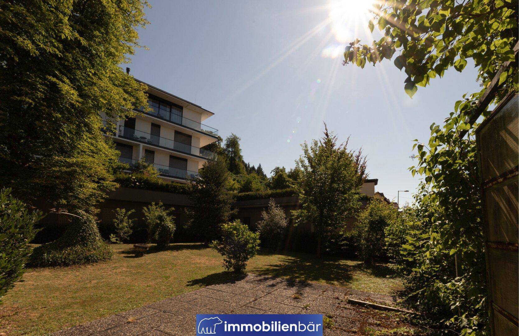 Sonniger Gartenbereich mit Rasenfläche, Bäumen und einem modernen Wohngebäude im Hintergrund.