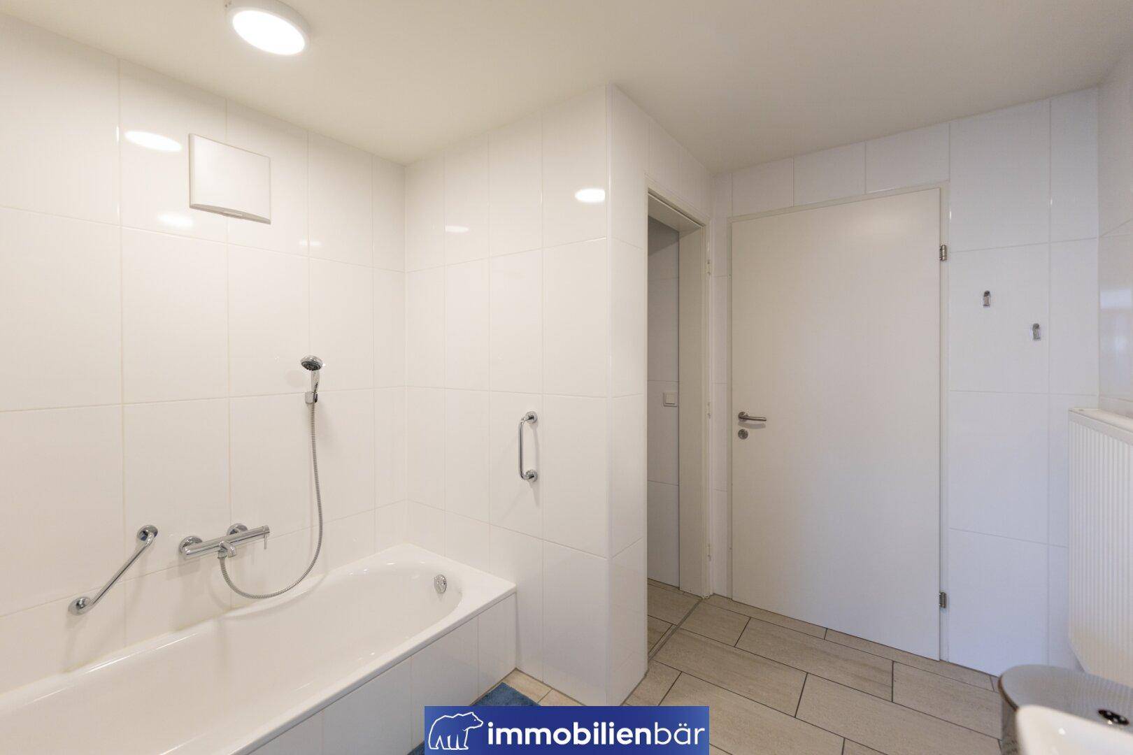 Weiß gefliestes Badezimmer mit Badewanne, Duscharmatur und einem Lüftungsventil an der Decke.