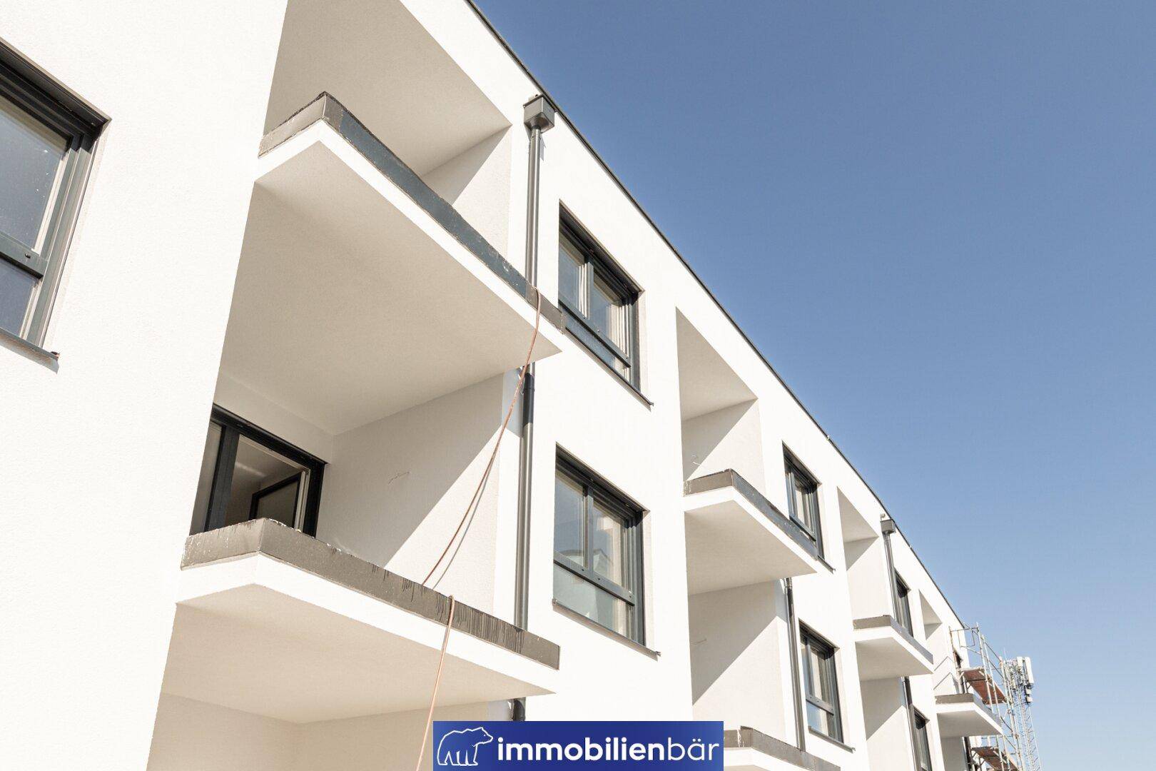 Moderne weiße Fassade eines Neubaus mit Balkonen unter klarem blauen Himmel.