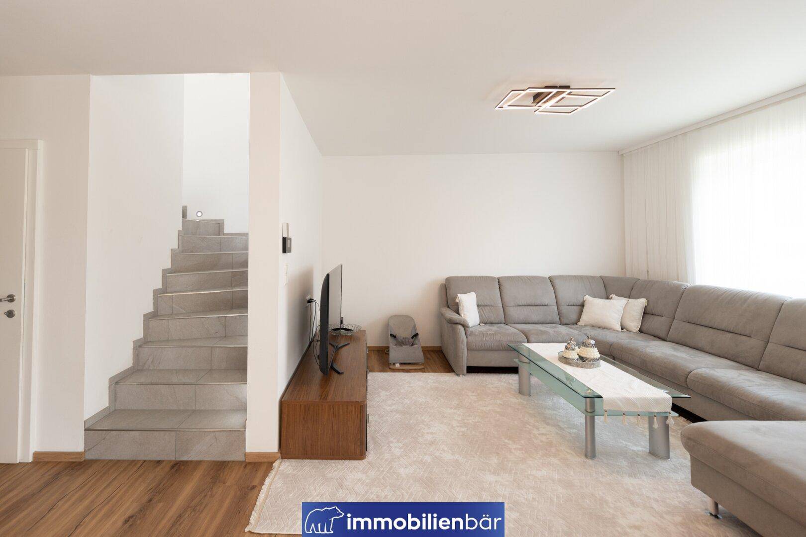 Helles Wohnzimmer mit großem Ecksofa, Fernseher und Treppe zum Obergeschoss.