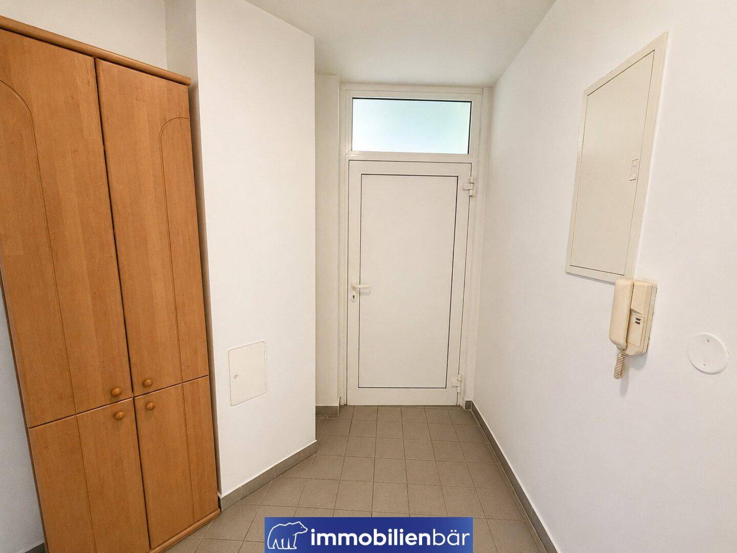 Eingangsbereich mit Fliesenboden, Holzschrank und heller Eingangstür, die viel Licht hereinlässt.