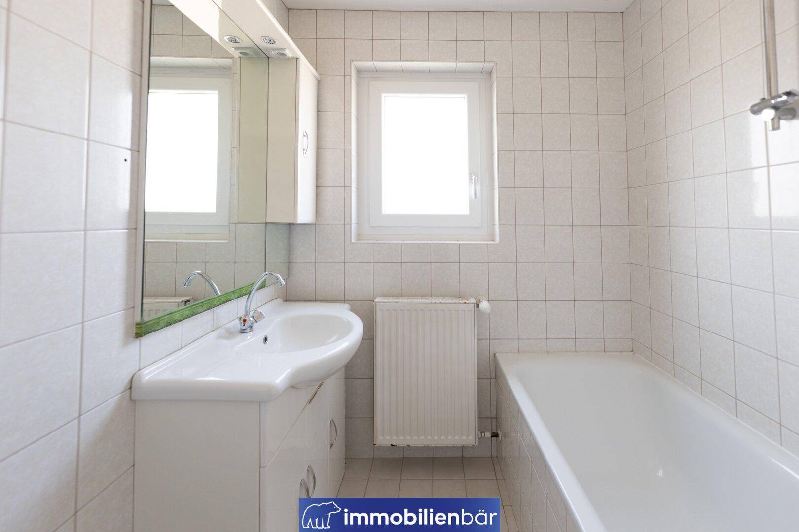 Helles Badezimmer mit weißen Fliesen, Badewanne, Waschbecken und Fenster.