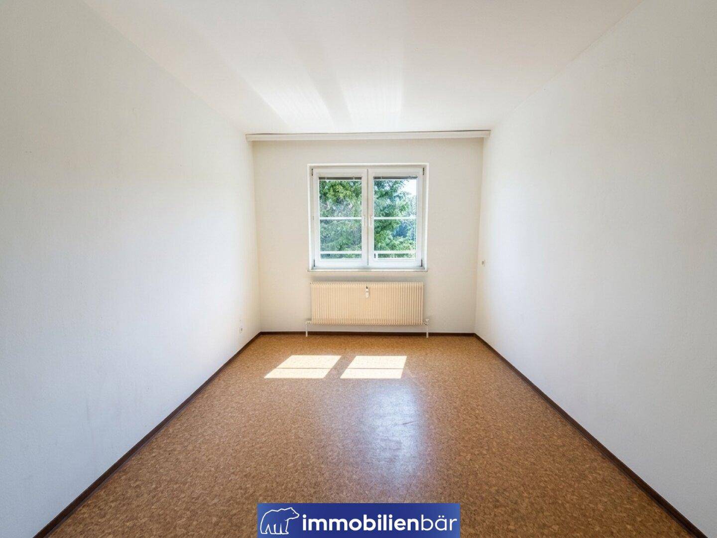 Helles, leeres Zimmer mit Korkboden und einem Fenster mit Blick ins Grüne.