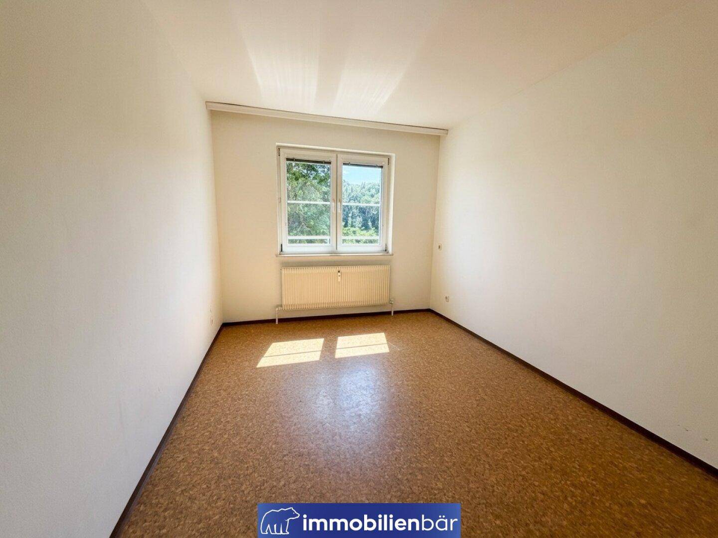 Unmöbliertes Zimmer mit Korkboden und einem Fenster, das viel Tageslicht hereinlässt.