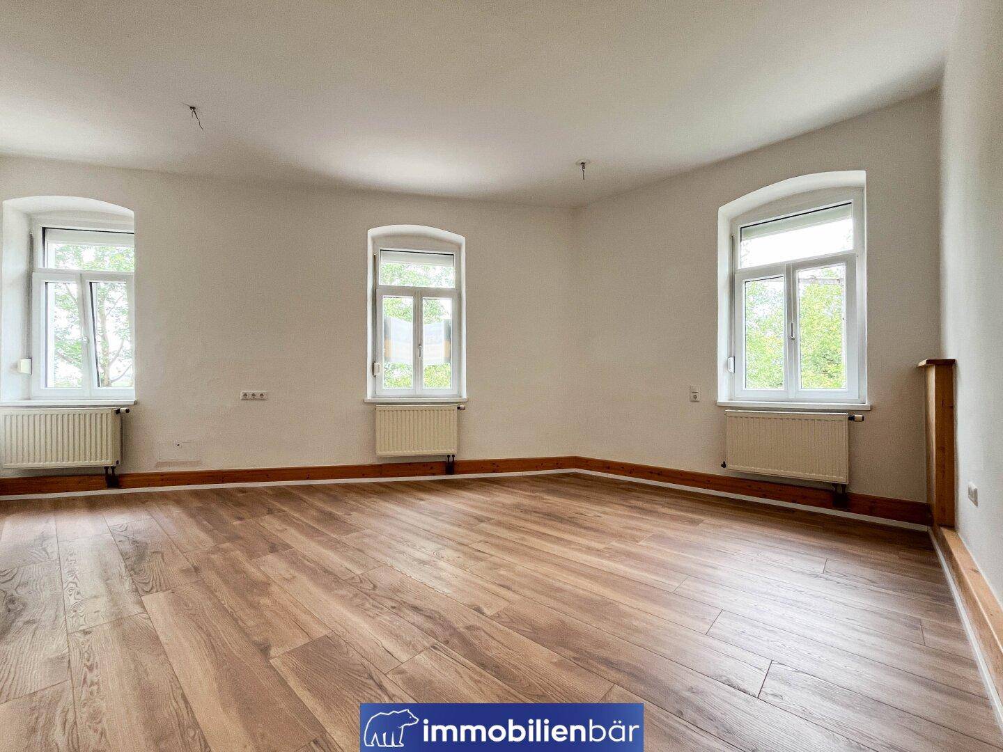 Geräumiges, helles Wohnzimmer mit drei großen Fenstern und ansprechendem Laminatboden.