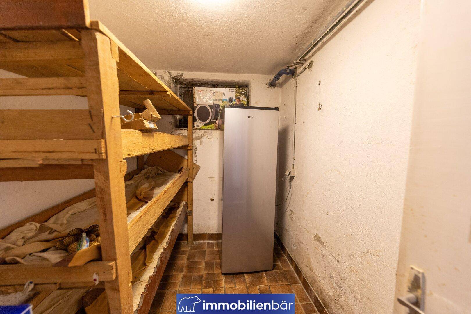 Kellerraum mit Holzregalen, einem Kühlschrank und rustikalem Fliesenboden für Lagerzwecke.