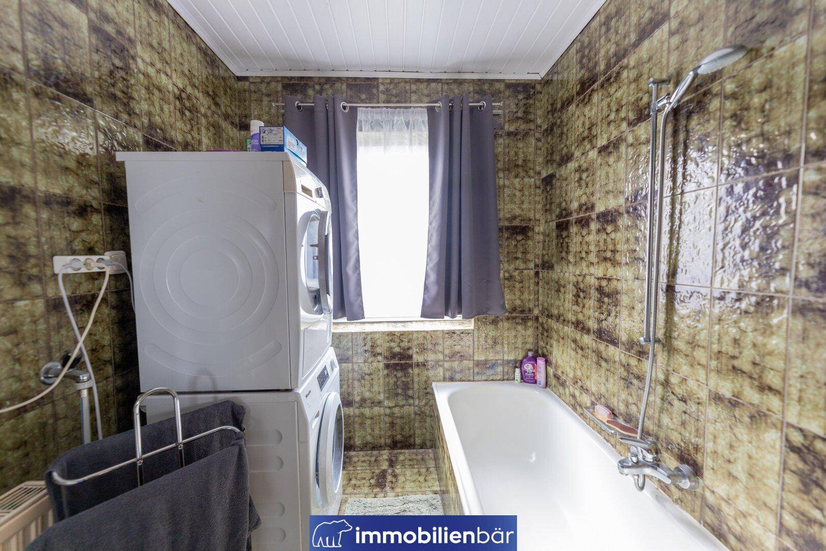 Badezimmer mit Badewanne, Waschmaschine und Trockner, gefliest in dunklen Farbtönen.