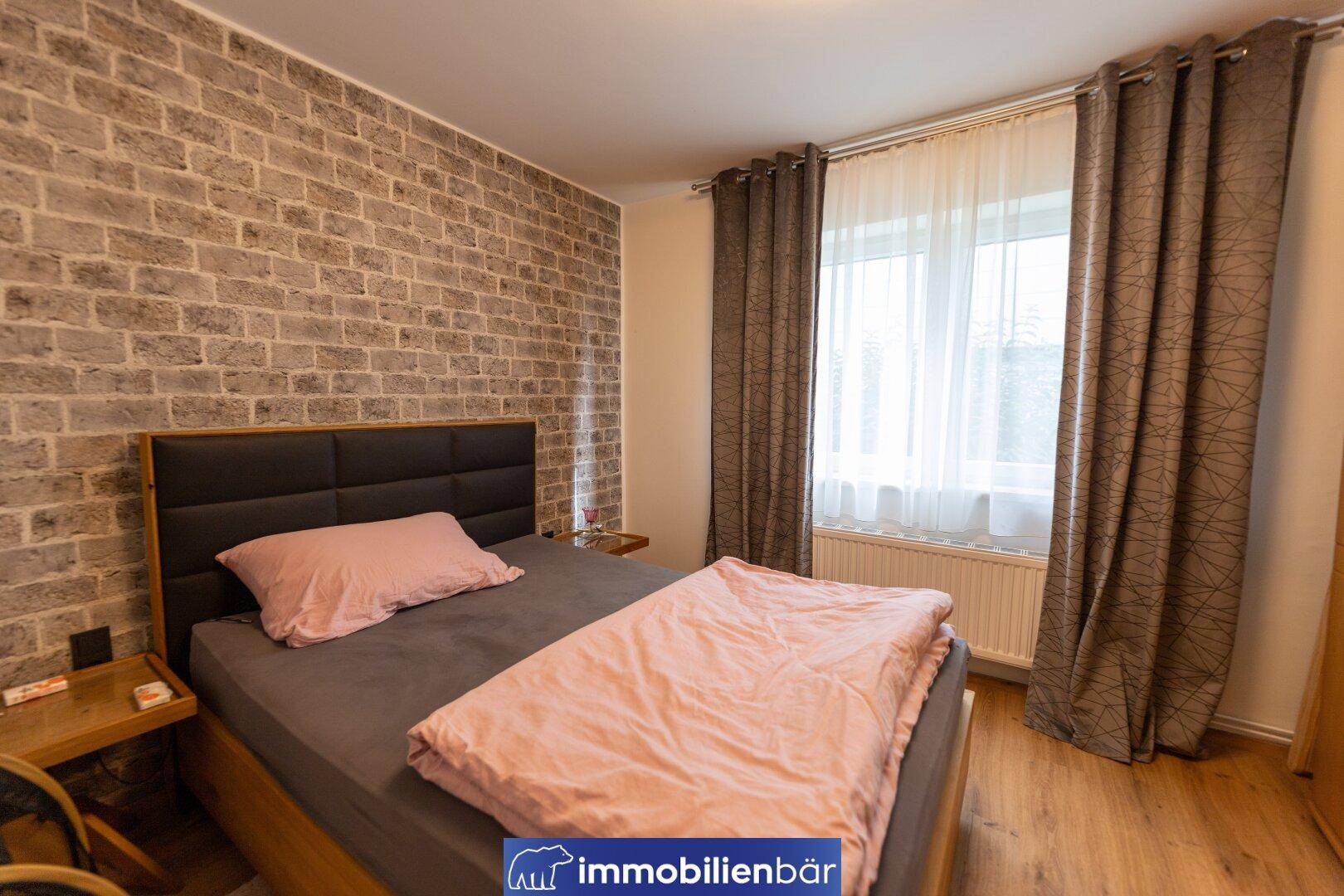 Schlafzimmer mit Ziegelwandakzent, Doppelbett und Fenster mit Vorhängen, bietet viel Tageslicht.
