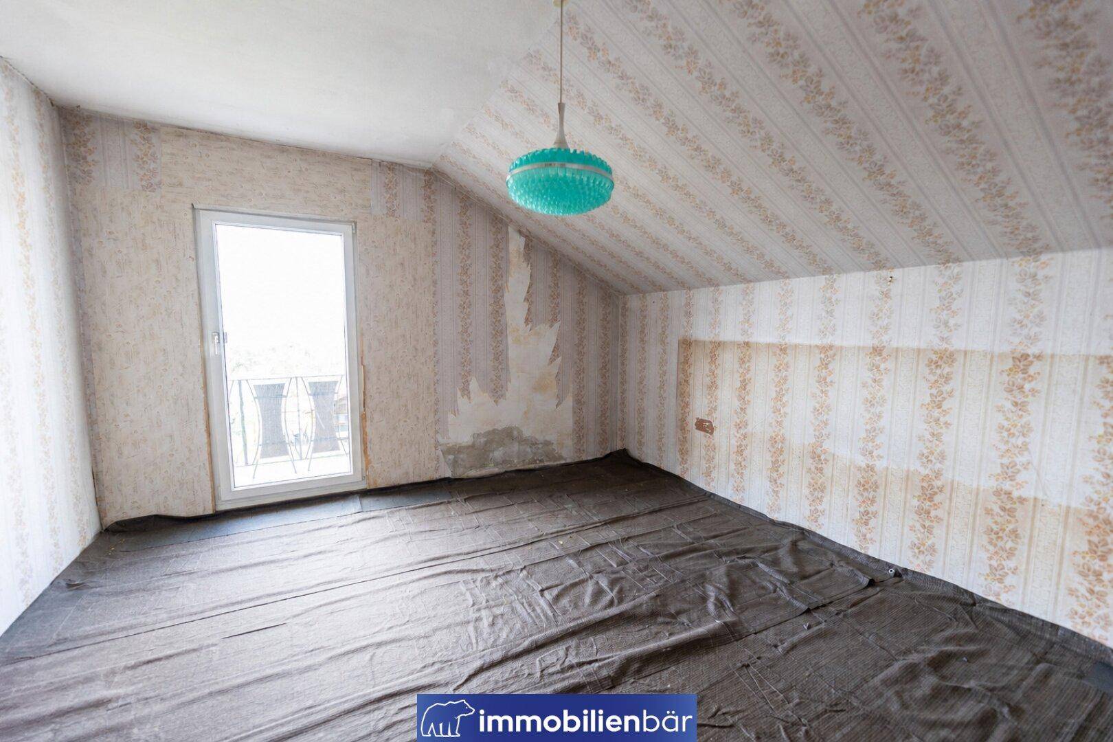 Leeres Dachzimmer mit schrägen Wänden, alter Blumentapete und Zugang zu einem französischen Balkon.