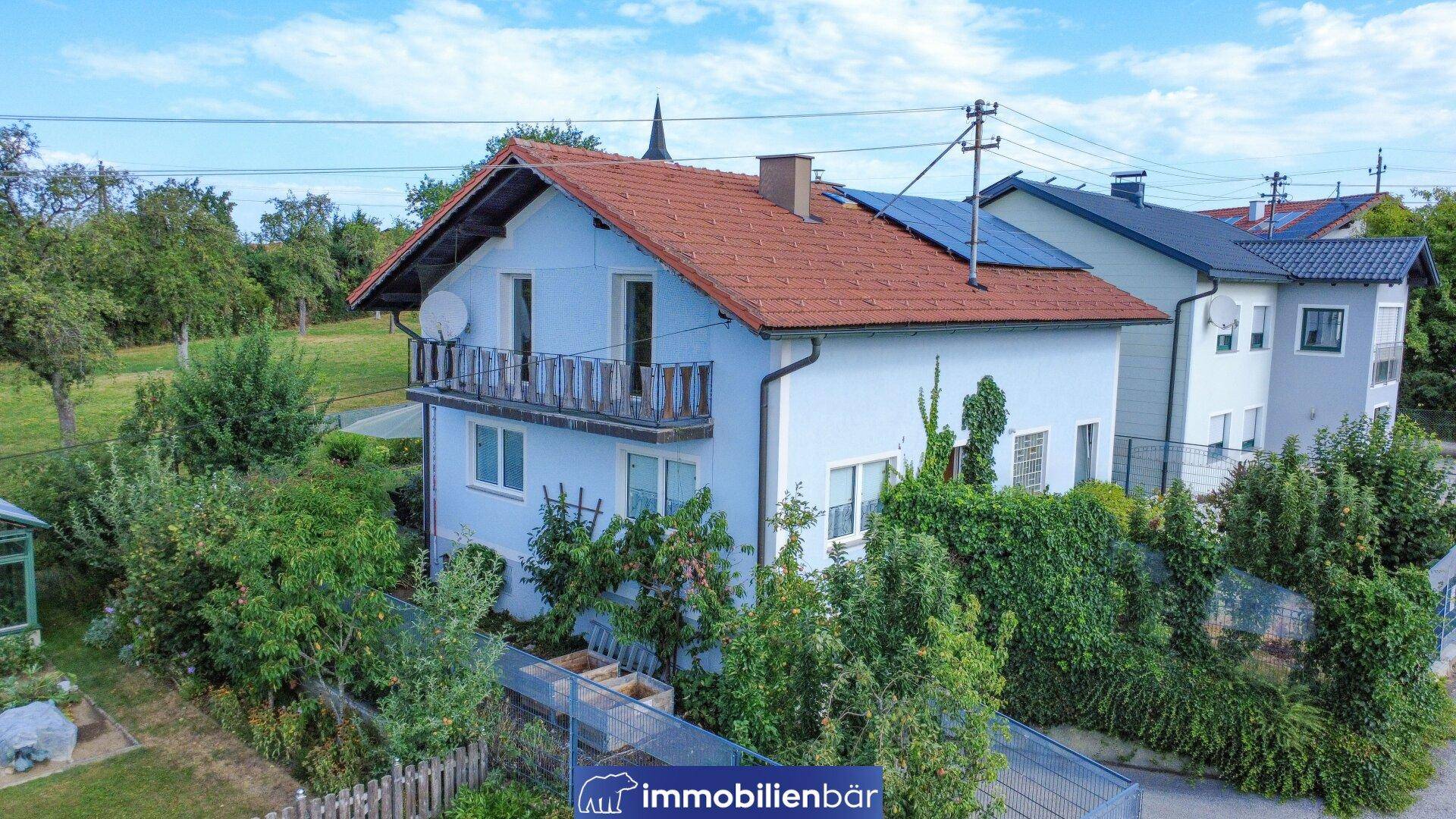 Seitenansicht des Hauses mit Balkon, umgeben von üppigem Grün und Solarpaneelen auf dem Dach.