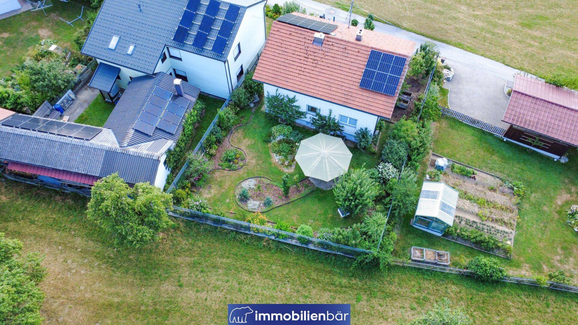 Vogelperspektive des Hauses mit großem Garten, Sitzbereich und angrenzenden Feldern unter blauem Himmel.