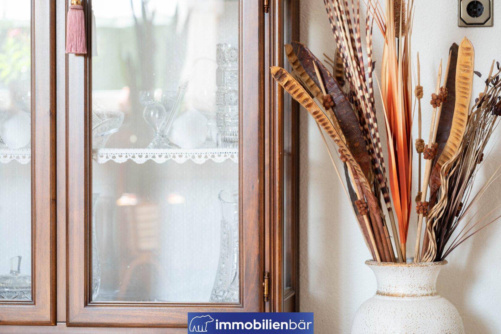 Detailaufnahme eines Holzschranks mit Glasvitrine und dekorativen Trockenblumenarrangements.