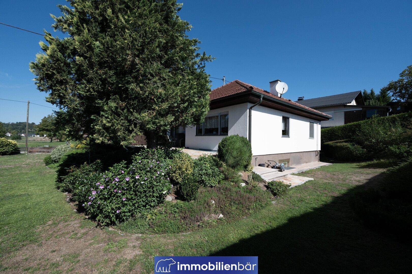 Seitenansicht des Hauses mit üppiger Bepflanzung und einem kleinen Gartenbereich unter blauem Himmel.