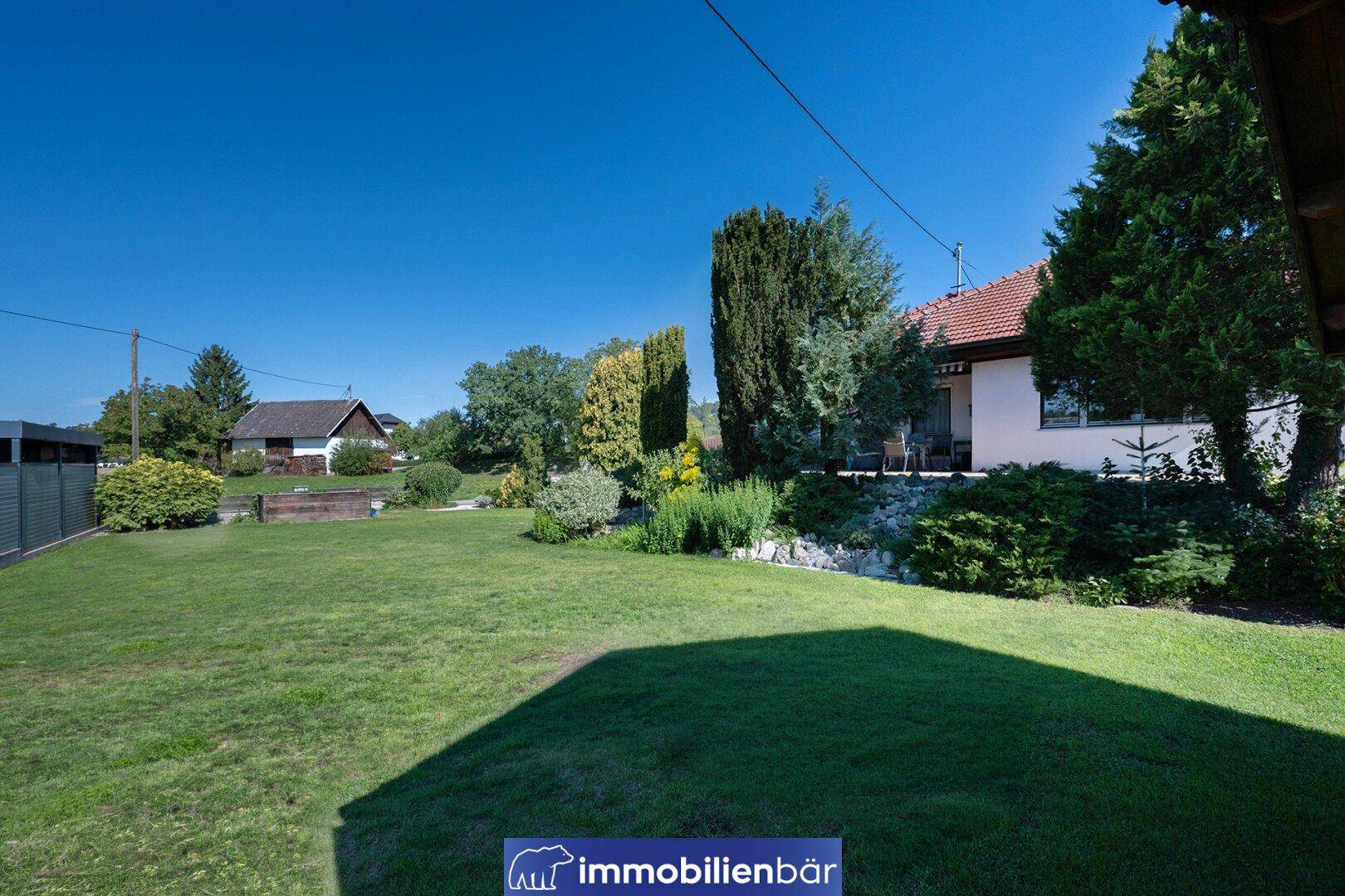 Sonnige Rasenfläche im Garten mit Blick auf die Rückseite des Hauses und die umliegende Natur.
