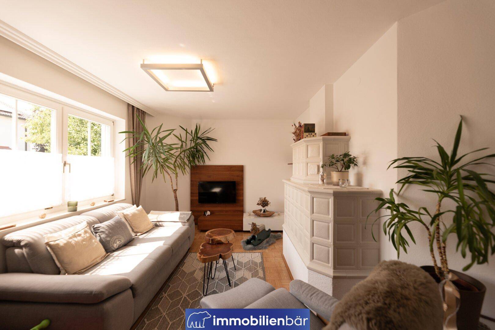 Helles Wohnzimmer mit großem Sofa, traditionellem Kachelofen und vielen Pflanzen.